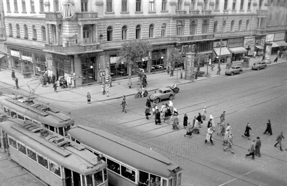 Sarokház a Nagykörút és a Rákóczi út kereszteződésében, a Blahán. Ezen az 1952-es képen még olvasható az EMKE felirat. A villamosok még mindkét irányban jártak, a zebrát még csak két pöttysor jelezte, nem volt se metró, se népszerűtlen árkádsor. Érdekes viszont a ház sarkán az első emelet magasságában kialakított, fedett forgalomirányító hely. Minket azonban persze a hirdetőoszlopok érdekelnek: ott van egyből kettő is. Túlélték a háborút, az államosítást - akkor épp az Állami Hirdető Vállalathoz tartoztak -, és mint látható, több fajtájuk is volt. 