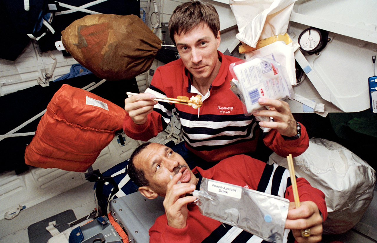 1994. február: az STS-60 küldetés egyik étkezési szünetében őszibarack-sárgabarack gyümölcslevet fogyaszt Bolden, miközben fölötte Krikaljev orosz űrhajós pálcikával falatozik.