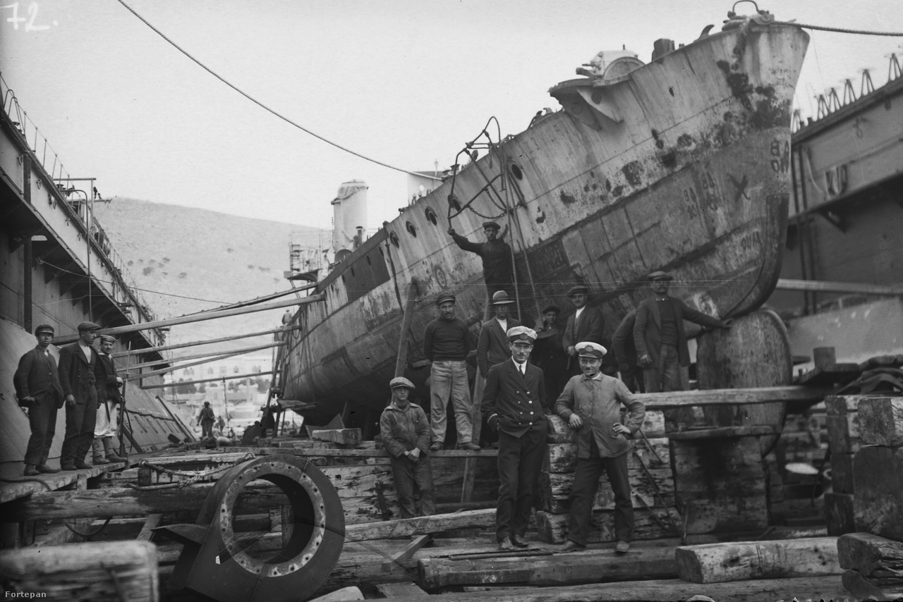 Csoportkép készülő hajóval. A kép a 20. század elején készülhetett, a bal szélen álló szuronyos puskás matróz, a munkások előtt álló hajóskapitány-szerű figura öltözéke is erre utal.