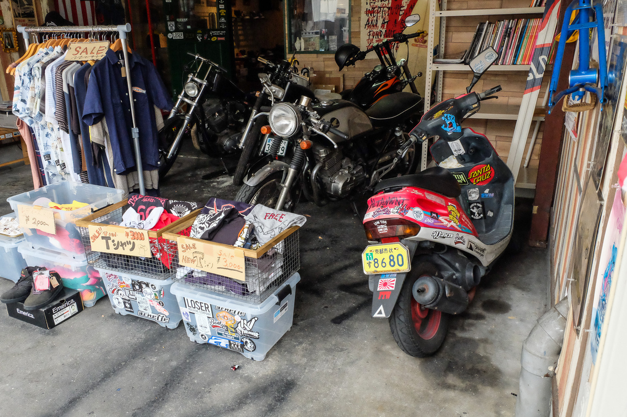 A helyhiány egészen vicces megoldásokra sarkallja a japánokat. Ez itt például egy vagány divatholmikat és rocker-relikviákat árusító, városszéli divatbolt. A tulaja egy harmincas, szőkített japán csávó, megkérdeztem, tőle, mit keresnek itt a motorok. Ja, mondta, a Harley az enyém, a két öreg japán gép a haverjaimé, akikkel motorozunk, a robogó a bolt futármotorja. Díszletnek pont jók ide, s így sikerült megoldtani a garázskérdést is