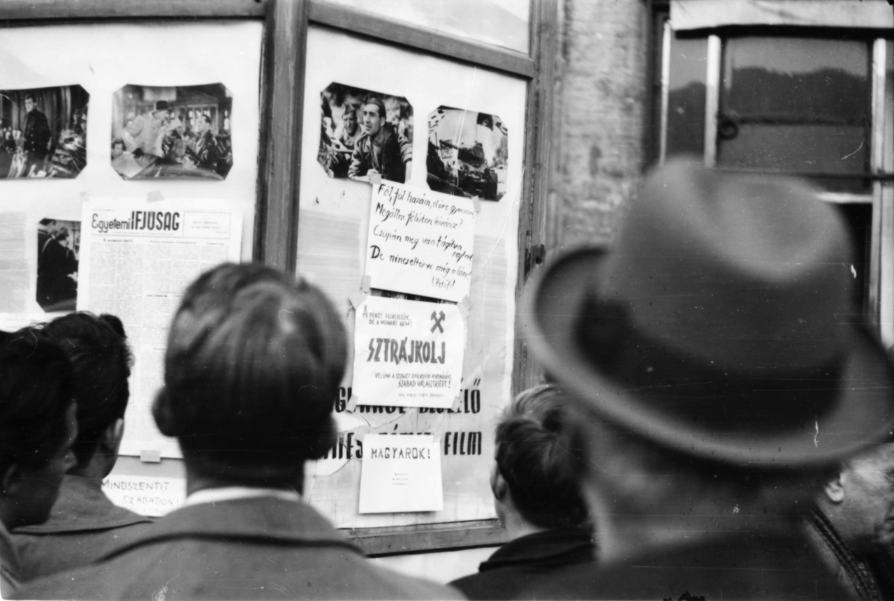 A korra jellemző üveg hirdetőoszlop. A mozihirdetések között, ha jól sejtjük, egy  keletnémet film, a Reichstag felgyújtásáról szóló Ördögi kör plakátja lenne látható, ha nem lenne átragasztva röplapokkal és az Egyetemi Újság friss számával. A kézzel írt buzdítások között van lelkesítő Petőfi-strófa, sztrájkfelhívás "a szovjet gyilkosok kivonásáért, a szabad választásokért", valamint egy rossz helyesírású követelés a hercegprímásért: "Mindszentit (...) szabadon!"