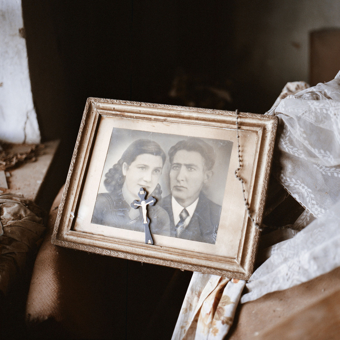 Házaspár portréja kereszttel és rózsafüzérrel egy üresen álló házból.