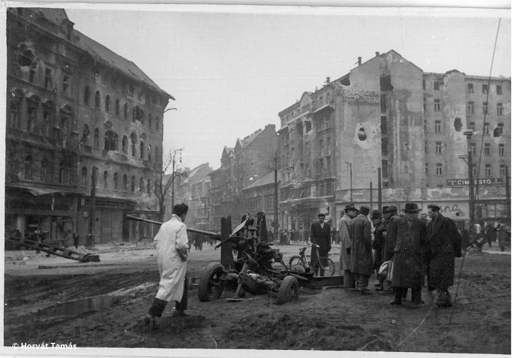 Ugyancsak a Baross tér, a lencse ezúttal a Rákóczi út felé néz. A felkelők november 3-án felállított ágyúját a Vörös Hadsereg hamar szétlőtte, a felkelők bázisa a Baross tér 19. számú háza még egy napig tartott ki.