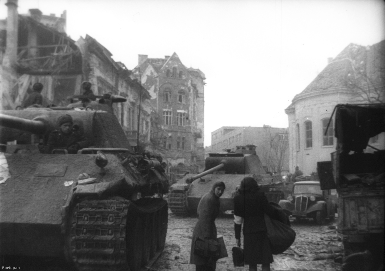 Ez a kép ugyanezt a Mészáros utcai helyszínt mutatja más
                        szögből. Két nő csomagokkal igyekszik valahova, a szovjet katonák a német harckocsikat vizsgálják. A mai utcaképet ide kattintva nézheti meg.