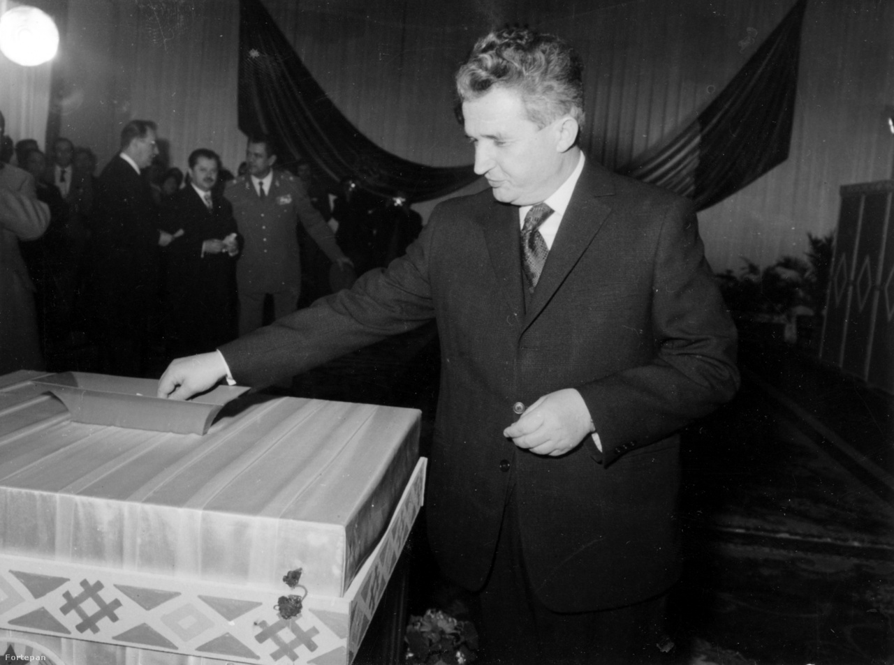 Nicolae Ceaușescut 1974-ben választották Románia első elnökévé. A kárpátok géniusza itt alighanem sajét magára szavaz. Ekkor már kilenc éve töltötte be a Román Kommunista Párt főtitkári posztját, az országban mind jobban elhatalmasodott a személyi kultusz. Az Új Élet Ceaușescu elnökké választását "történelmünk nagy pillanatának" nevezte, és a kor divatjának megfelelően nagy összeállításban emlékezett meg a pártvezér életéről. A kép hátterében Manea Manescu román miniszterelnök, jobbra mellette Fazekas János erdélyi magyar politikus, a Román Kommunista Párt Politikai Végrehajtó Bizottságának tagja. Ceaușescut 1989-ben kivégezték. A Ceaușescunál két évvel idősebb Manescu felesége Ceaușescu lánya, Maria volt. Manescu 2009-ben halt meg Bukarestben. Az újságíróként és történészként is ismert Fazekas János Sütő András szerint magas állami és párttisztségeiben (vagy azok ellenére) sokat tett az erdélyi magyarságért. 2004-ben Budapesten halt meg, az MTA megbízásából kutatott a magyar fővárosban.