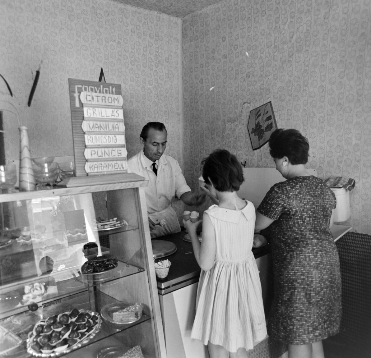 1968 – CukrászdaOrosházán vagyunk, egy cukrászdában. Kicsike az üzlet, ez abból látszik, hogy a kézmosási lehetőséget csak nagy ügyeskedés árán sikerült beszorítani a pult mellé: a víztartály alján csap, alatta pedig tálca a piszkos víz felfogására. Ez csak a KÖJÁL (= Közegészségügyi és Járványügyi Állomás) előírásainak betartása miatt kellett, nyilván ragaszkodtak hozzá, hogy ahol élelmiszer-fogyasztás történik, ott kézmosási lehetőségnek kell lennie. (És vajon vécé volt?) A fagylaltkínálat mai szemmel nézve bizony egyoldalú, nincsen semmilyen gyümölcsös (a citromosat nem lehet annak nevezni), a felsorolt fagylaltok ugyanannak a tejszínes alapnak különböző ízesítései. Ettől még finomak lehettek, én a rumosdiót választottam volna. A „grillás” szónak „s”-sel való írása általános volt (ahogy a „masszás” szónak is, bár a franciás „grillázs”, „masszázs” helyesebbnek számított). A víztartály tetején csíkos kéztörlő.