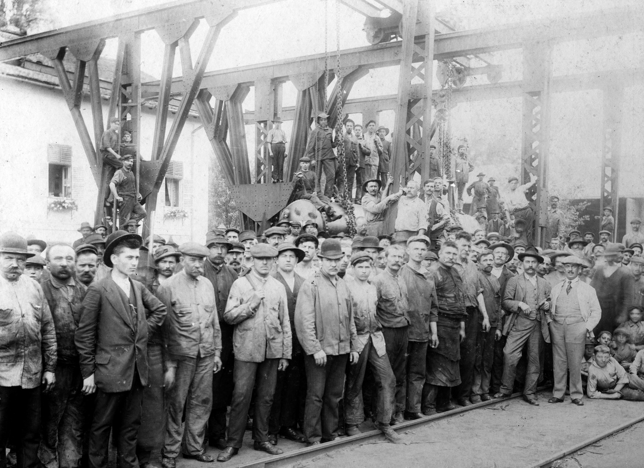 1902 – MunkásgyűlésEzeknek a hajógyári munkásoknak, úgy látszik, nem mondta a fényképész, hogy „Barátságos arcot kérek”, nyilván nem is akartak barátságosnak látszani. Talán szakszervezeti megmozdulás, talán sztrájkra készülnek – ekkor már erősödőben volt a munkásmozgalom. A jobb szélen gyerkőcök, ők persze vidámabbak: könyököl a legelső a földön, mint majd teszi tizenvalahány év múlva a hazaküldött katonafotókon. A gyerekek előtt áll egy világos ruhás, köpcösebb férfi zsebre dugott kézzel: ő lehet a gyáregység vezetője (vagy a tulajdonos?), aki az iménti beszédében megpróbálta csitítani a munkásokat. A legérdekesebb a baloldalt elöl álló sötét ruhás fiatalember, feltolt keménykalapban. Ő a szakszervezeti vezető, később majd illegális kommunista lesz, ha megölik, utcát és laktanyát fognak elnevezni róla. Nyilván derék, szimpatikus ember, de az ilyeneknek az emlékét úgy összenyálazták később a pártpropagandisták, hogy csak lenéző ellenszenvvel tudtunk rájuk gondolni. Ha pedig életben marad, pártvezető lesz, ami kiöli belőle ezt a távolba néző, eltökélt, rokonszenves fiatalembert.