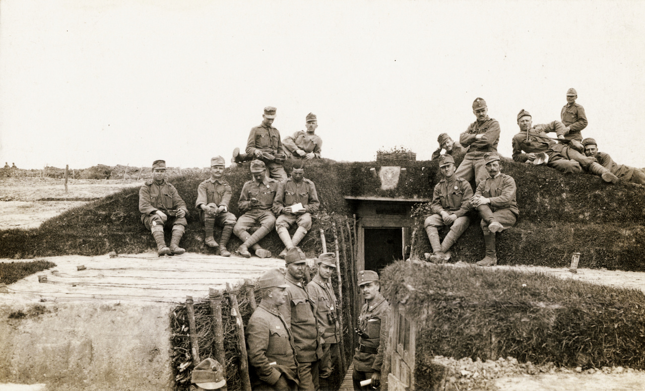 1915 – LövészárokZajlik a Nagy Háború, de ez még nem a rémes része. Lám, milyen takaros kis lövészárkot csináltak a fiúk! Elégedettek is a munkájukkal, tűrik az okvetetlenkedő fényképészt. Fönt a dombocskán a jellegzetes könyöklő „katonafotós” tartást látjuk, méghozzá automatikusan a kép középvonalára szimmetrikusan könyökölnek a bal, illetve jobb könyökükre. A lejjebb ülők közül az egyik nem figyel, sapka sincs rajta, ejnye, fiam! Levelet olvas, persze, a menyasszonyától vagy az ifjú feleségétől – pedig úgy látszik, nemrég vannak még kint a fronton, lesz ez még ígyebb is. A mellette ülő társa meg mobilozik – oppardon, nem mobilozik, mert ekkor még nem volt mobil. De akkor mit csinál, hiszen az egész test- és kéztartása olyan? A zsebóráját vizsgálgatja elmélyülten?