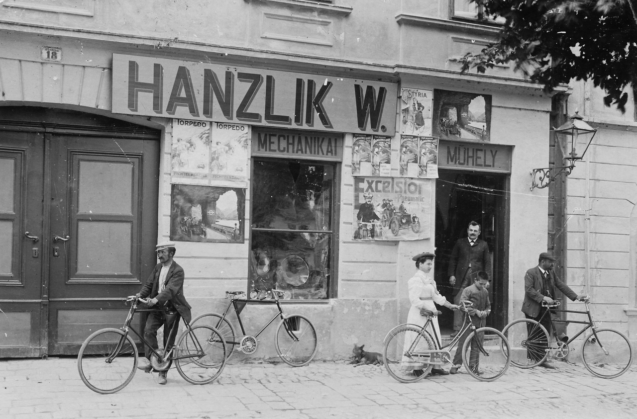 1913 – Vasparipa Az ajtóban, gondolom, maga Hanzlik úr áll, a tulajdonos, nézi, ahogyan a boldog vevők szanaszét hordják a frissen vásárolt kerékpárokat. De az is lehet, hogy csak beugrottak hozzá biciklin valami alkatrészért. Mindenesetre a nő kezében női vázas kerékpár van, míg a többi mind férfigép, szóval kicsit reklámízű a felvétel. Fájdalmas belegondolni, hogy egy ilyen kis helyen, mint Magyaróvár (mely csak 1939-ben egyesült Mosonnal) naprakész „mechanikai műhely" létezett, amely az éppen forgalomba került nyugati dolgokat is forgalmazta, mint az a plakátokból kiderül. Hová zuhant mindez a második háború után! A műhely a Fő utca 18.-ban működött, Hanzlik úr német lehetett, erre keresztnevének „W” betűjéből következtethetünk, de a vezetékneve olyan általános keletosztrák-nyugatmagyar-északhorvát-délszlovák, hogy azt a Jóisten se bogozza ki. Ha Bécsben élt volna, Hanslicknak írta volna, Pozsonyban meg Hanzlíknak. Testtartásán látszik a támogató jóindulat, mellyel kuncsaftjait szemléli. Isten áldja a tisztes ipart!