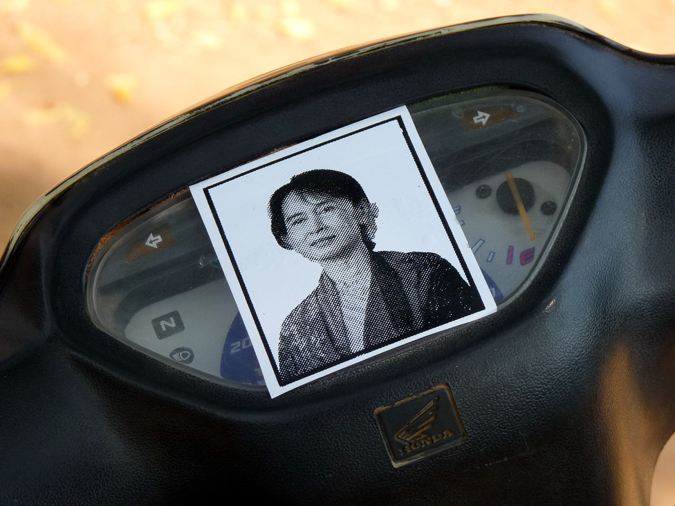 Változó idők. A robogón Ang Szan Szú Kji ellenzéki vezető fotója. A Nobel-békedíjas politikusnő hosszú időt töltött a katonai kormányzat börtöneiben és házi őrizetben. 1990-ben betiltott pártjának működését néhány éve engedélyezték újra, és a 2010-es választáson a parlamentbe is bejutott. Ang Szan Szú Kji a diktatúrával szembeni ellenállás vezetője és szimbóluma, korábban egy ilyen kép nyilvános mutogatásáért még letartóztatták volna a tulajdonosát. 
