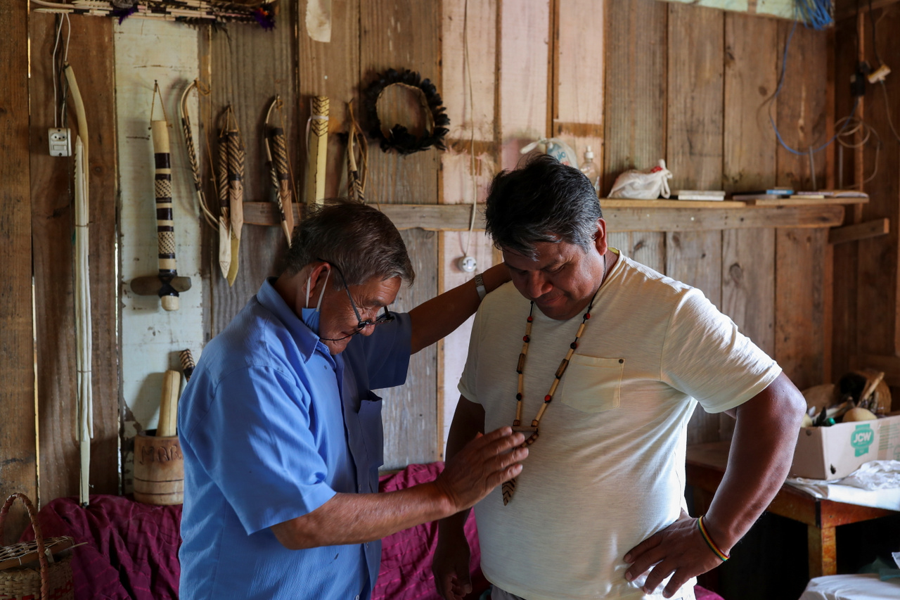Vile Ndille (78), a törzs egyik időse felesége által kézzel készített nyakláncot ajándékoz a falu vezetőjének, Lazaro Kamlemnek. A kép bal oldalán a háttérben kézzel készített íjak, nyilak, lándzsák és egyéb eszközök figyelhetők meg.