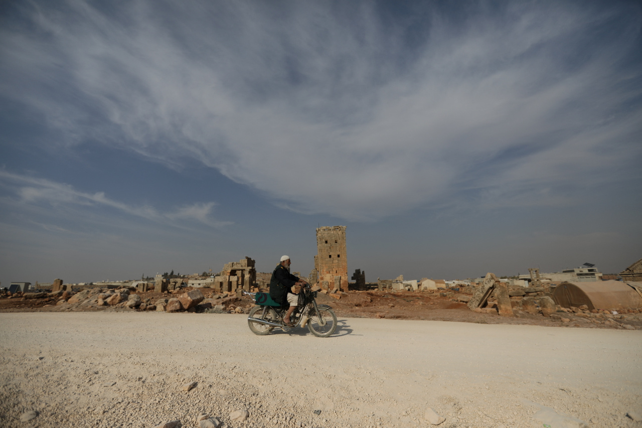 Férfi motorozik a sarjablei romok előtt. A kép jobb oldalán láthatók a kövek között felállított sátrak, melyekben a családok élnek.