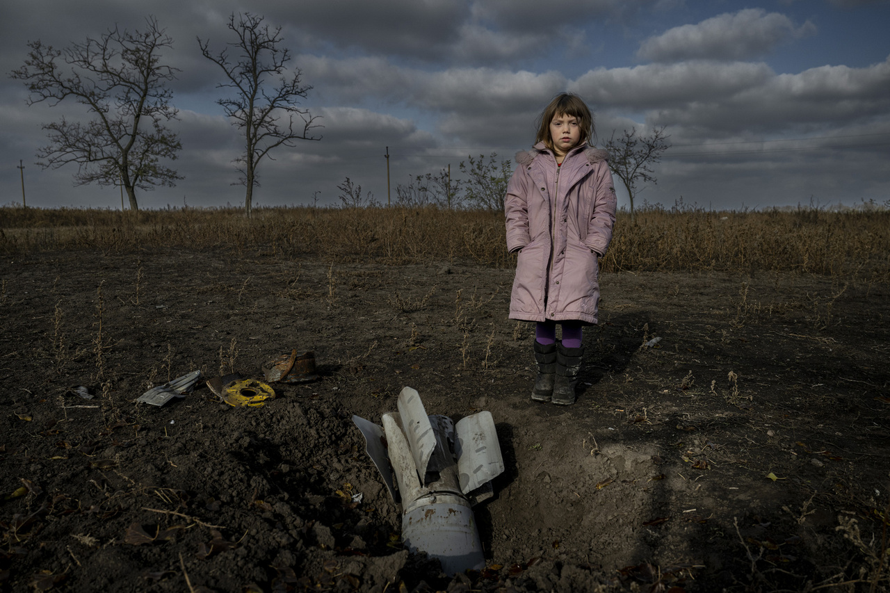 Emberábrázolás, portré (egyedi) 2. díj: A pokol kertje (Biljaivka, Herszon régió, Ukrajna, 2022.11.10) - Mariia (9 éves) az ukrajnai Herszon régióban található Biljaivkában él édesanyjával és nővérével.
                        A család nem menekült el az orosz megszállás elől; ennek fő oka a pénzhiány és az volt, hogy nem volt kihez menniük. Hét hónapot töltöttek orosz megszállás alatt, és a pincéjükben bujkáltak a légitámadások alatt.
                        Mintegy száz orosz katonát telepítettek Biljaivkára, emiatt a falubeliek többsége a biztonságosabb szomszédos falvakba menekült. Biljaivkában nem volt sem áram, sem folyóvíz; és a mobiltelefon-szolgáltatás is akadozott. Mariia életét a puszta szerencse mentette meg, éppen akkor ment be a kültéri vécéjükbe, amikor egy rakéta becsapódott a kertjük végébe. „Nagyon megijedtem, amikor becsapódott a rakéta, nem gondoltam semmire, csak elestem. Remegett a föld és a ház is. Anyukám nem enged a kertben játszani, mert még mindig ott vannak a kazettás bombák maradványai mindenhol, amelyek felrobbanhatnak, ha rájuk lépsz."
                        