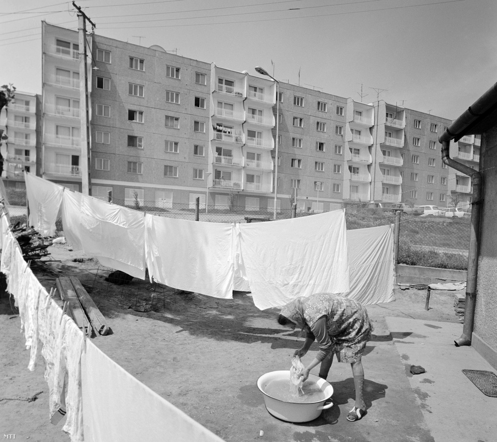 1977. május. Mosott ruhákat tereget egy asszony a paksi atomerőmű dolgozóinak épült új lakótelep szomszédságában. A nagyközség 10 ezres lakossága húsz év alatt megkétszereződött. Paks 1979-ben kapott ismét városi címet. (Ezt még 1871-ben vesztette el, a mezővárosi rang megszüntetésével.)
                        Új gondolat volt, hogy az építőmunkásokat nem barakkokban helyezik el, hanem már ők is új panelházakba költözhettek, amelyek később az üzemeltetőknek adtak otthont. Több mint kétezer lakás épült így. Az új panelházakhoz kapcsolódik az úgynevezett "tulipános vita" is: néhány panelházat vörös-fehér ívelt mintákkal díszítettek. "Az egész ország szürkében lakik, miért legyenek a paksiak különbek? Mi lesz, ha mindenki ilyet akar?" - kérdezte az akkori építésügyi miniszter, leállítva a kezdeményezést.  