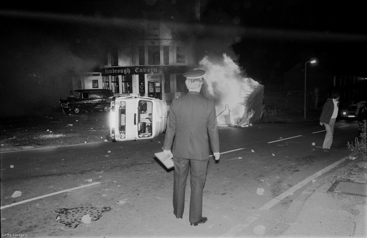 Southall, Anglia, 1981. július 4. Rendőr áll egy felborult rendőrségi furgon és egy égő teherautó előtt, az ázsiaiak és a szkinhedek közt dúló küzdelmek után