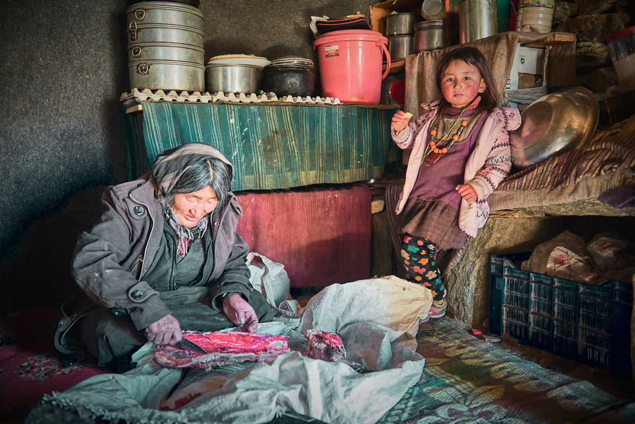 Changpa nagymama jak húst darabol az ebédhez. Mellette kis unokája majszol egy darab kenyeret (Ladakh, 2018. február)