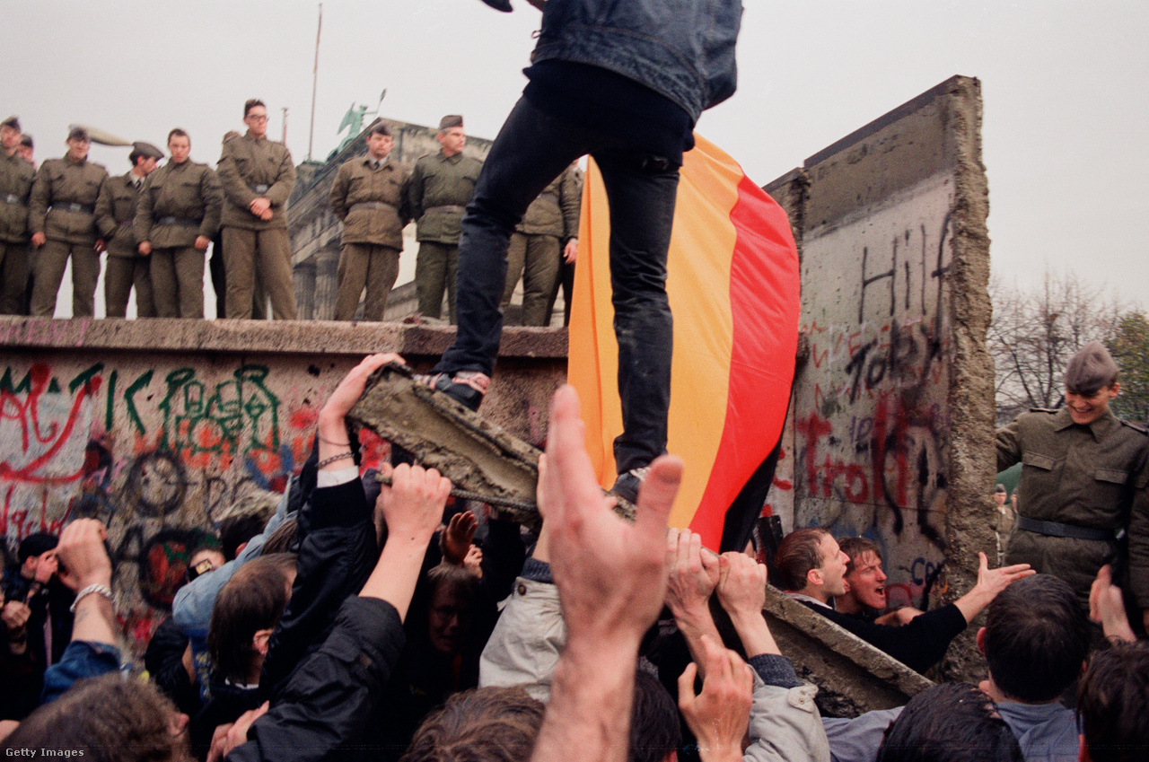 Berlin, Németország, 1989. november 10. Egy férfi a berlini fal egy ledöntött darabján áll tüntetőtársai segítségével, kezében német zászlóval, miközben kelet-német határőrök figyelik