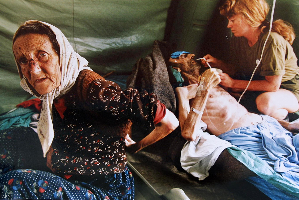 Ez a kép 1995 július 11-én készült. Az idős nő és a hosszú idő óta éhező férje megsérültek a szerbek srebrenica elleni támadásaiban.
                        Akik tudtak, a még nem lerombolt bosnyák városokba menekültek a hadsereg kegyetlensége elől. Srebrenica lakossága ezért egy-két év alatt kilenc ezerről 42 ezerre duzzadt. A városban már kezdetektől ostromkörülmények uralkodtak: a legtöbb menekült az utcán, bármiféle fedezék nélkül húzta meg magát, miközben folyamatosa volt szerbek tüzérségi támadása, alig volt víz és áramot is csak a házilag buherált generátorok termeltek. Az emberek éheztek, mindennapos volt az erőszak, virágzott a prostitúció és a fekete piac. 1993-ban Srebrenicában a járt az ENSZ jugoszláviai békefenntartóinak (UNPROFOR) a vezetője, a francia Philippe Morillon és egy lakossági gyűlésen azt mondta, nem kell aggódni, a város az ENSZ védelme alatt áll és az ENSZ sosem fogja cserben hagyni a srebrenciaiakat.